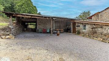 Un bâtiment rustique en pierre, avec une allée de gravier, des bancs en bois et un espace abrité comprenant une table et des chaises.