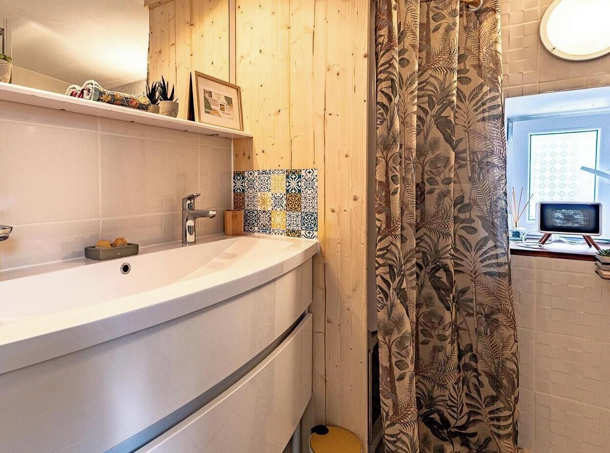 Een badkamer met een wit wastafelmeubel, een spiegel, een wandpaneel van hout, een douchegordijn met een patroon en een klein raam.
