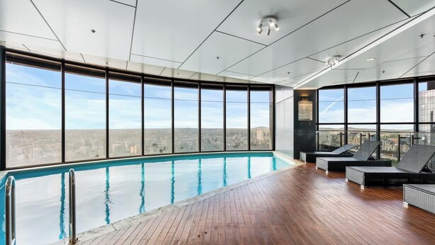 Piscina interior moderna con vistas a la ciudad, terraza de madera y zona de descanso.