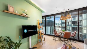 Una sala de estar moderna con un televisor de pantalla plana, un estante de madera con objetos decorativos, una estantería escalonada, una zona de comedor con sillas y una mesa, y ventanas amplias que ofrecen vistas a la ciudad.
