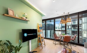 Ein modernes Wohnzimmer mit Flachbildfernseher, einer Holzhälfte mit Dekoration, einem Himmelsregal, einem Essbereich mit Stühlen und Tisch sowie großen Fenstern mit Blick auf die Stadt.