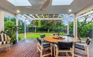 Eine überdachte Terrasse mit einem runden Holztisch und Stühlen, einem Deckenventilator und Blick auf einen Garten.