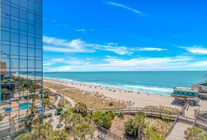 A beach view with a boardwalk, a pool area, and a building with a glass facade. - Yachtsman S703A Studio (Myrtle Beach)
