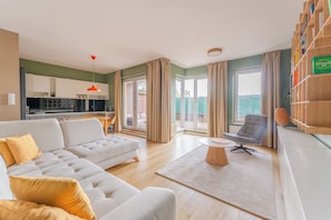 Ein modernes Wohnzimmer mit einer Couch, einem Couchtisch und einem Sessel. Es gibt eine Küchenzeile mit Schränken und einem Esstisch. Große Fenster mit Vorhängen sind zu sehen.