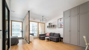 Uma sala de estar moderna com um sofá, uma mesinha e uma cadeira. Há um guarda-roupa grande e uma janela com persianas.