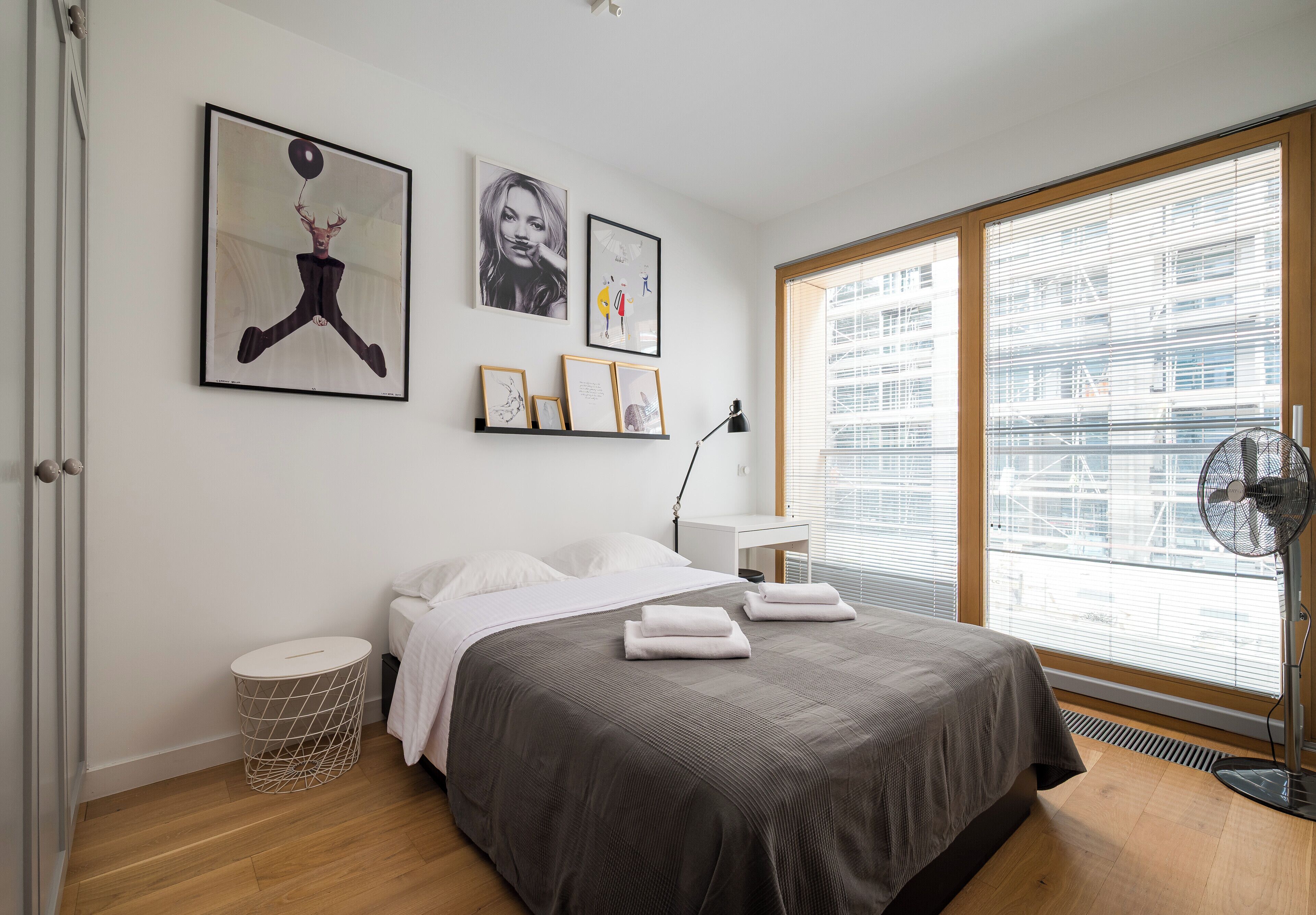 Un dormitorio con cama, mesita de noche, ventilador de pie y ventana con persianas.