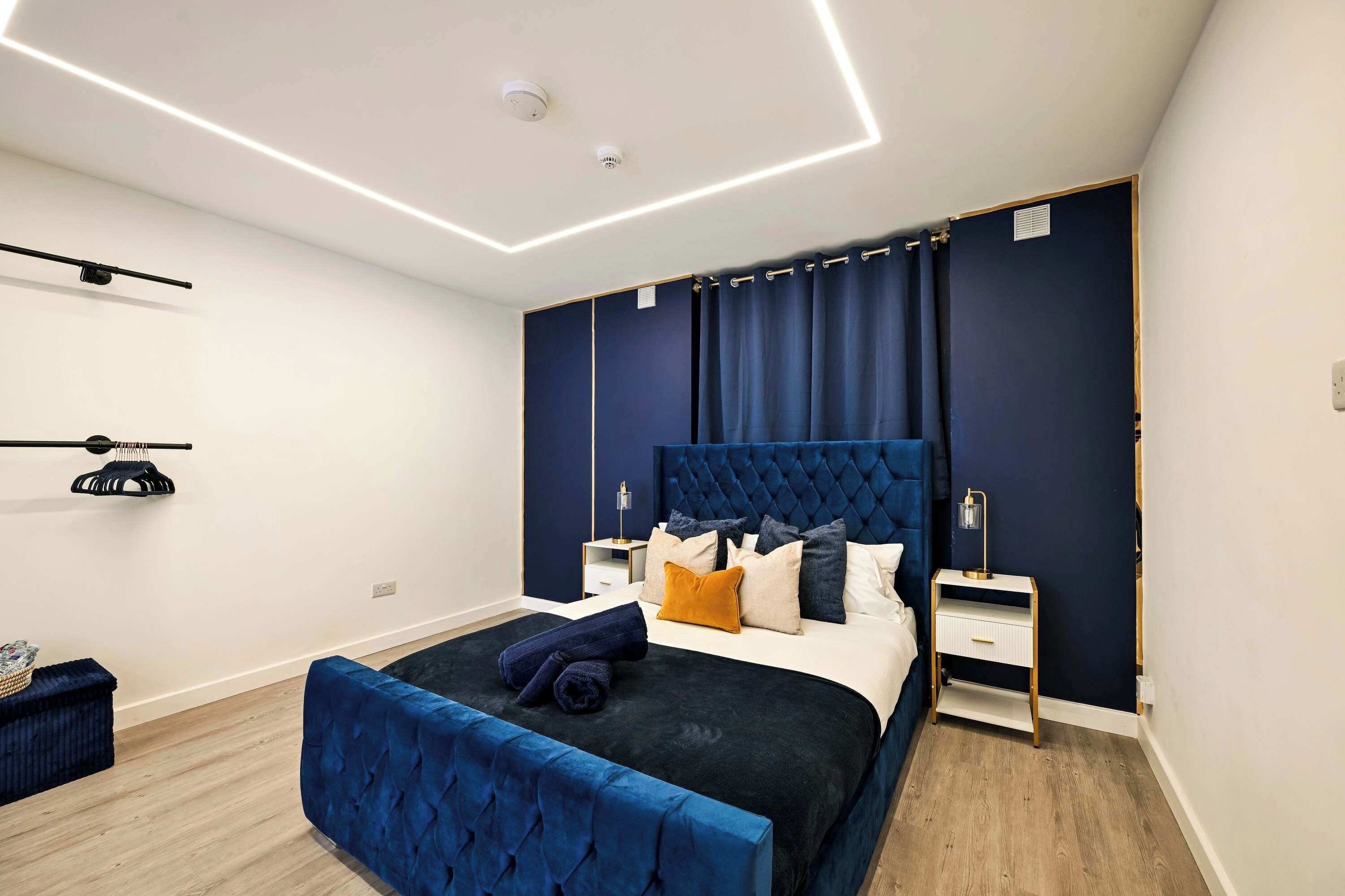 Ein modernes Schlafzimmer mit einer gepolsterten Kopfstütze, einem großen Bett mit vielen Kissen, einem Nachttisch mit Lampe und einer blauen Sitzbank.