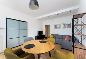 Ein modernes Wohnzimmer mit einem runden Holztisch, grünen und grauen Sofas, einer schwarzen TV-Konsole und einem Metallregal.