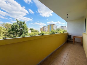 Ein Balkon mit gelben Wänden, gefliestem Boden, einem kleinen Tisch, einem Stuhl und Blick auf Gebäude und Bäume.