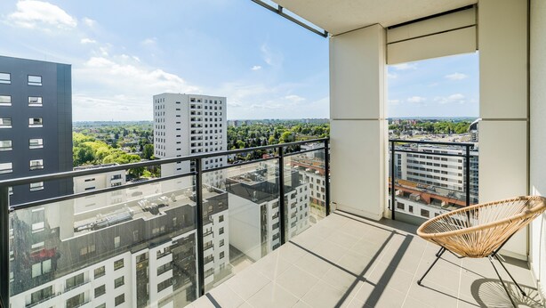A balcony with a view of surrounding buildings, a wicker chair, and a tiled floor. - Lasek Marceliński 12th Floor Studio (Poznań)