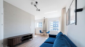 A modern living room with a blue sofa, a wooden entertainment unit, a dining area with two chairs, and a large window with a city view. - Lasek Marceliński 12th Floor Studio (Poznań)
