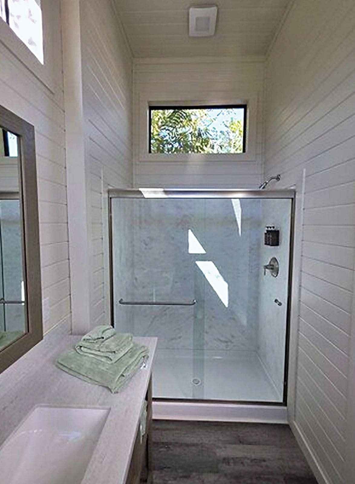 A narrow bathroom with a glass shower enclosure, a sink with a towel, and a window above the shower.
