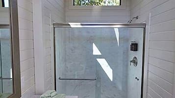 A narrow bathroom with a glass shower enclosure, a sink with a towel, and a window above the shower.