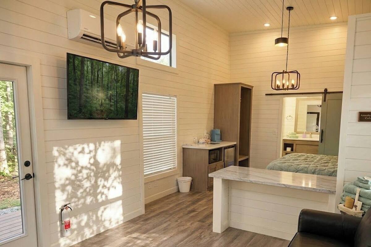A compact, well-lit room with a wooden floor, a large window, a painting, a kitchenette, a bed, and a mirror.