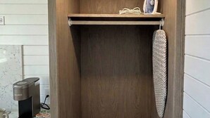 A compact, wooden closet with a shelf and hanging space, located in a room with white paneled walls. - Camp Cottonwood (Cottonwood)