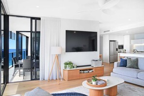 Ein modernes Wohnzimmer mit großem Flachbildfernseher, einem Holztisch, einem grauen Sofa und einem Balkon mit Gartenmöbeln.
