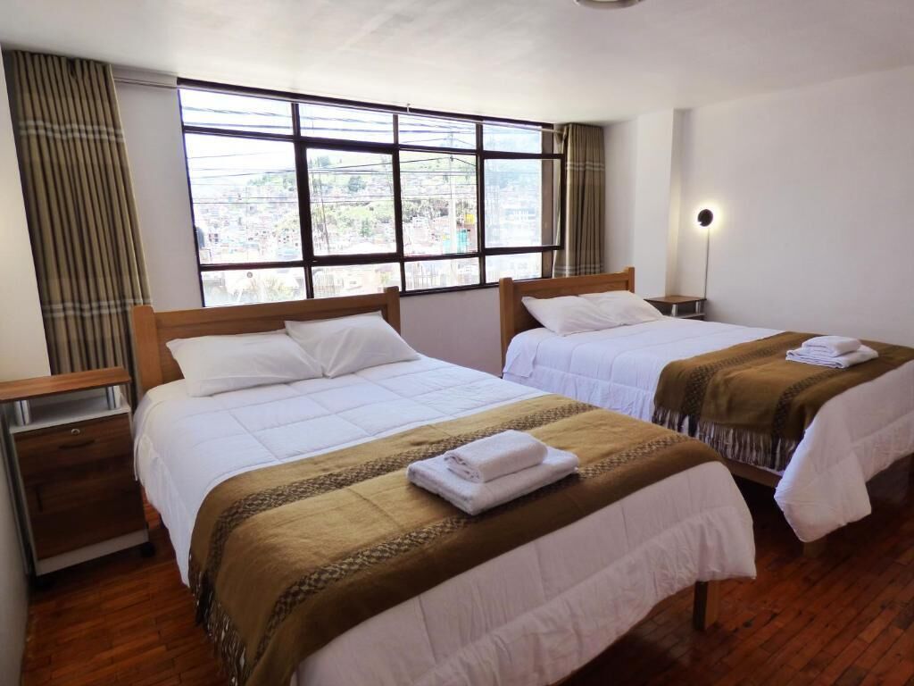 A hotel room with two beds, a large window, a bedside lamp, and a view of the city.
