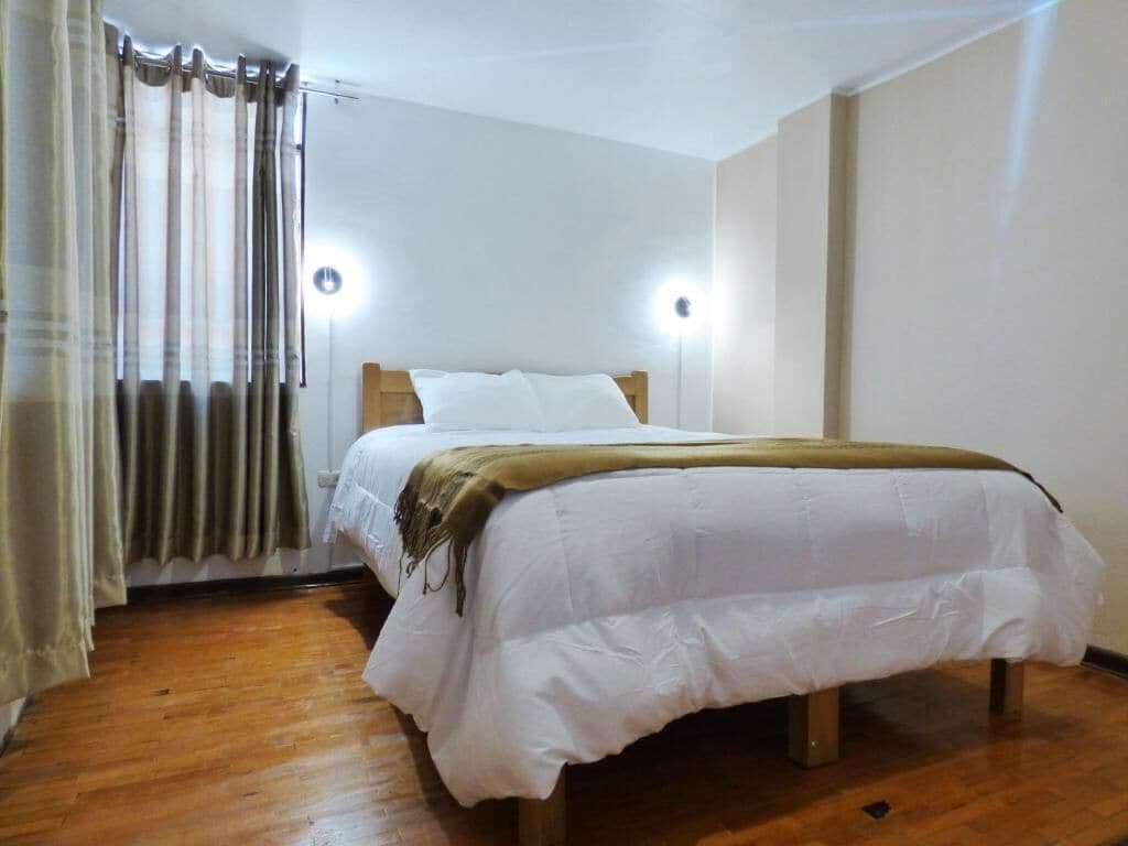 A single bed with a wooden frame, white bedding, and a brown throw blanket. There are two wall-mounted lights on either side of the bed.