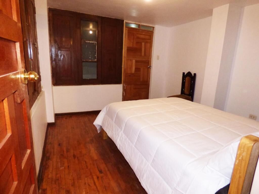 A bedroom with a wooden floor, a bed with a white bedspread, a wooden door, and a window with a view of the outside.
