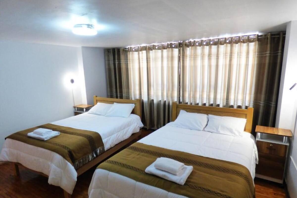 Habitación de hotel con dos camas, cabeceros de madera, una mesita de noche con lámpara y una ventana con cortinas.