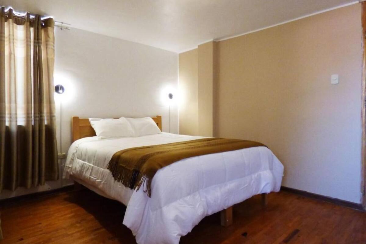 Una cama individual con edredón blanco y manta marrón, cabecera de madera y dos lámparas fijadas a la pared.