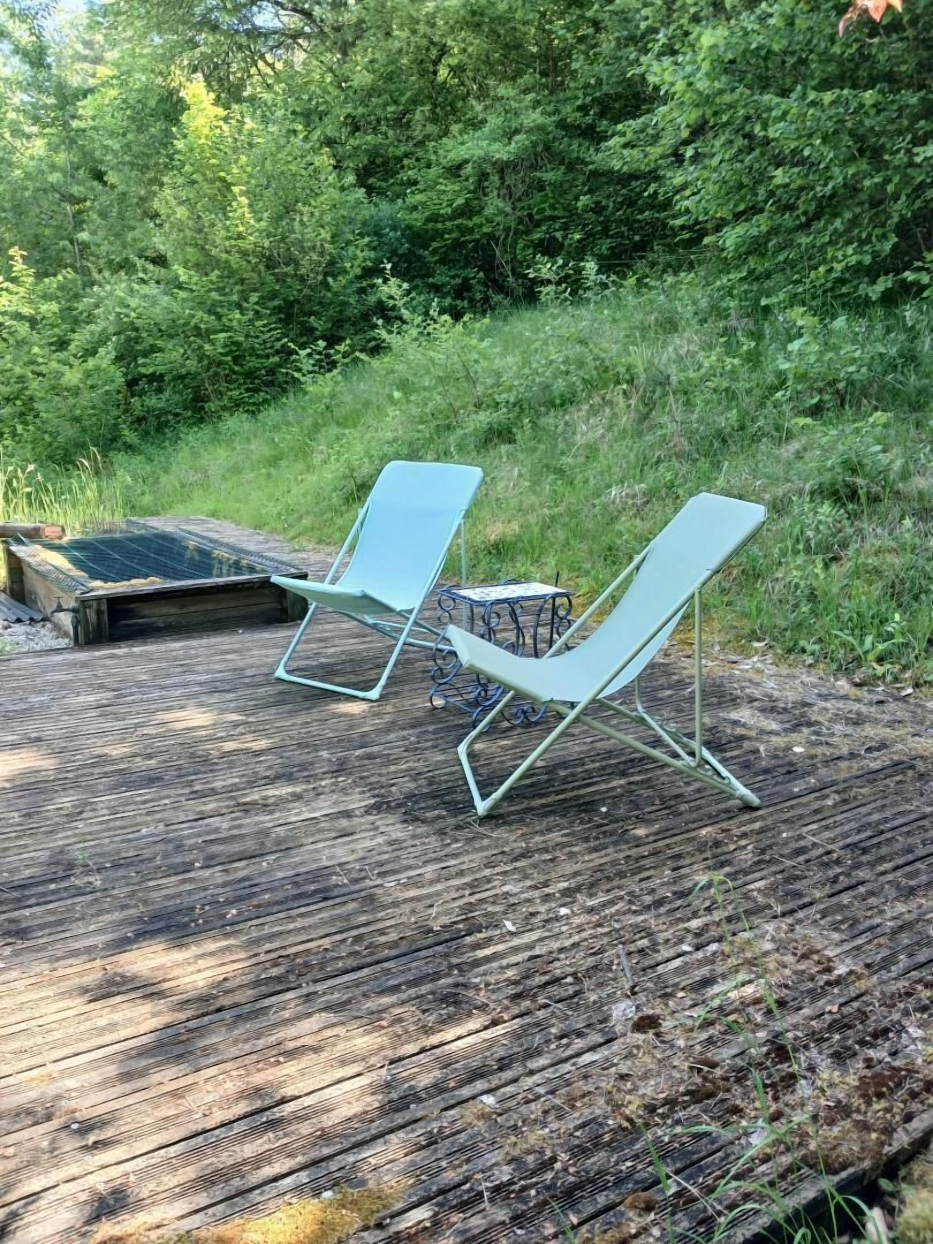 Two light blue lounge chairs on a wooden deck with a metal table, surrounded by dense greenery.