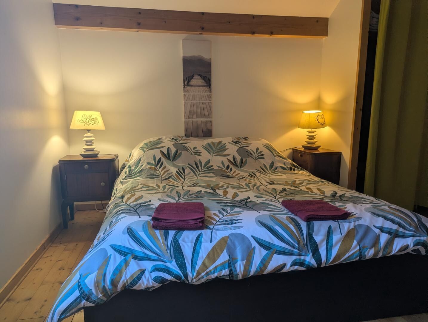 A bedroom with a bed, two bedside tables, a lamp on each table, a wooden headboard, and a wall-mounted artwork.