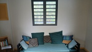 Ein Zimmer mit Holzboden, einem Fenster mit Jalousien, einer Couch mit Kissen und einem kleinen Holztisch mit Büchern.