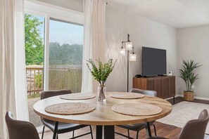 Ein Essbereich mit einem runden Holztisch, vier Stühlen, einer Vase mit Grünpflanzen, einer Stehlampe, einem Flachbildfernseher und einem Schrank.