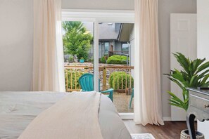 Ein Schlafzimmer mit einem großen Bett, Blick auf einen Balkon mit Stühlen und Pflanzen sowie einer Tür rechts.