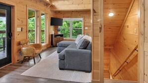 L’intérieur d’un chalet accueillant, avec un canapé gris, des murs en bois, un téléviseur à écran plat et une grande fenêtre.