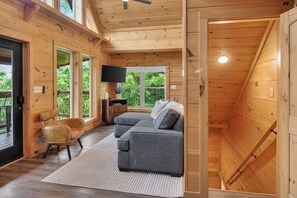 Een gezellig interieur van een blokhut met een grijze bank, houten wanden, een flatscreen-tv en een groot raam.