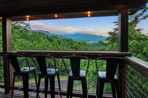 Um deck de madeira com cadeiras de metal, com vista para uma paisagem verde exuberante e montanhas ao longe.