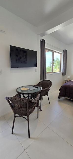 Ein Zimmer mit einem an der Wand befestigten Fernseher, einem Esstisch mit Stühlen, einem Bett mit violetter Bettdecke und einem Fenster mit Vorhängen.