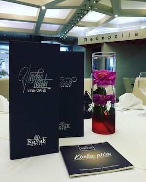 A reception area with a reception desk, a black card with 'Novak Hotel' branding, a glass vase with purple flowers, and a menu card. - Novak Hotel (Zvornik)
