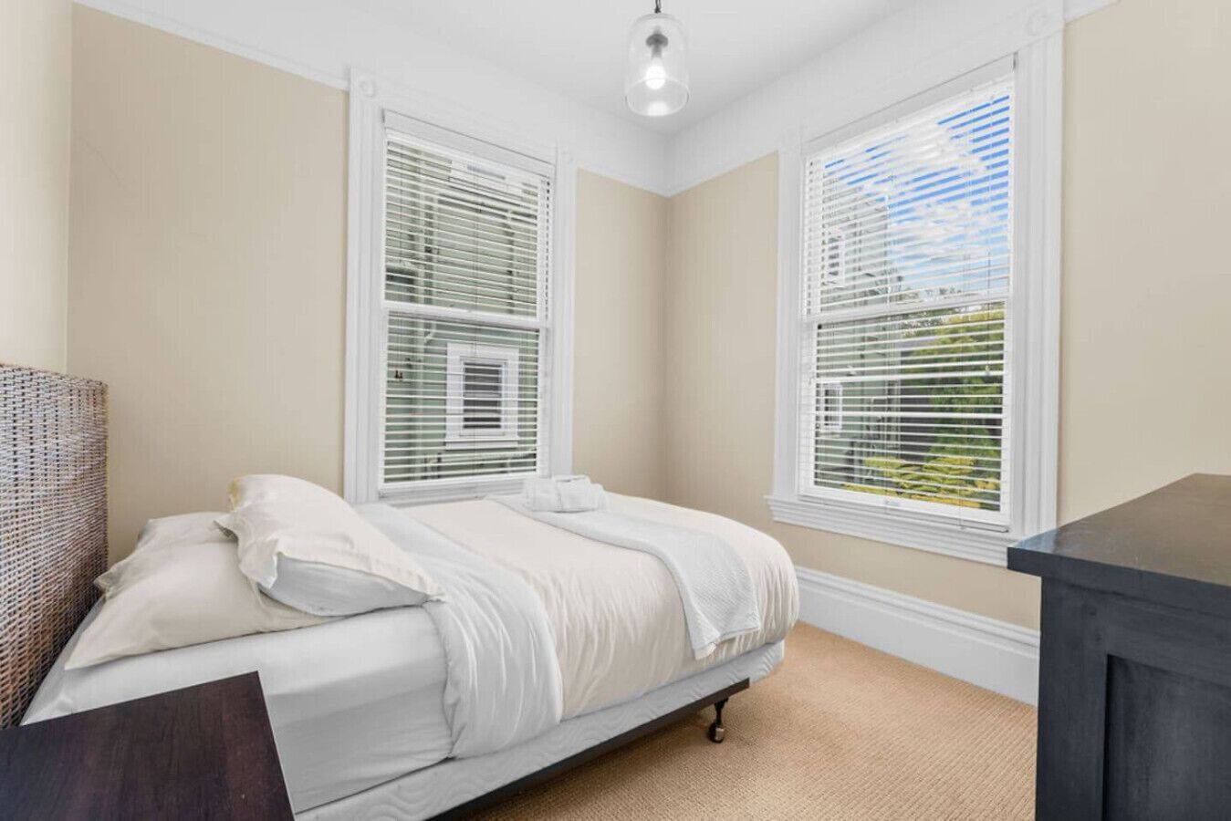 A bedroom with a bed, two windows with blinds, a ceiling light, and a dark piece of furniture.