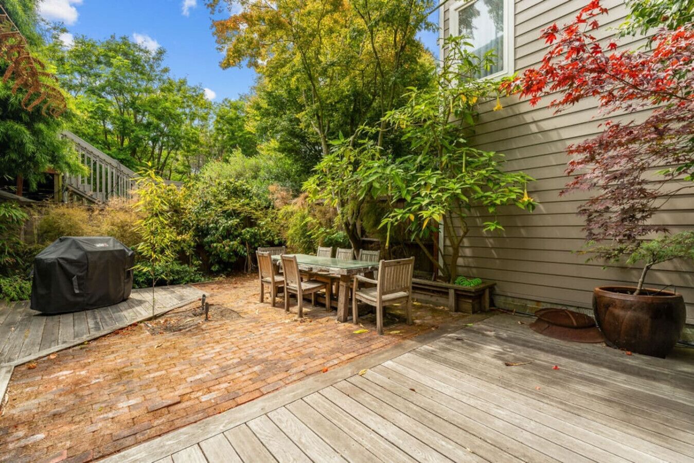 Een houten terras met een tafel en stoelen, omgeven door weelderige begroeiing en een overdekte barbecue.