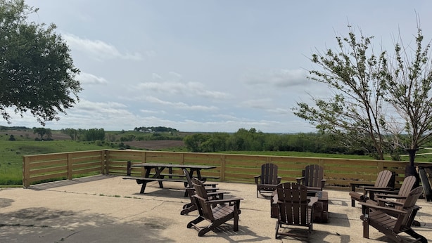 An outdoor seating area with picnic tables and Adirondack chairs, surrounded by a wooden fence and a view of a green field.