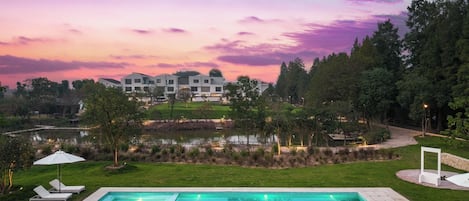 A swimming pool with lounge chairs, a pond, and a building in the background.