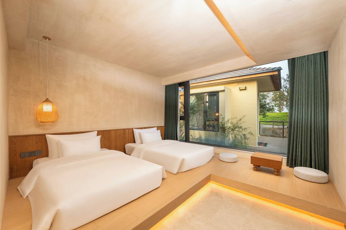 Una habitación de hotel moderna con dos camas, un banco de madera, una mesita pequeña y un ventanal con cortinas.