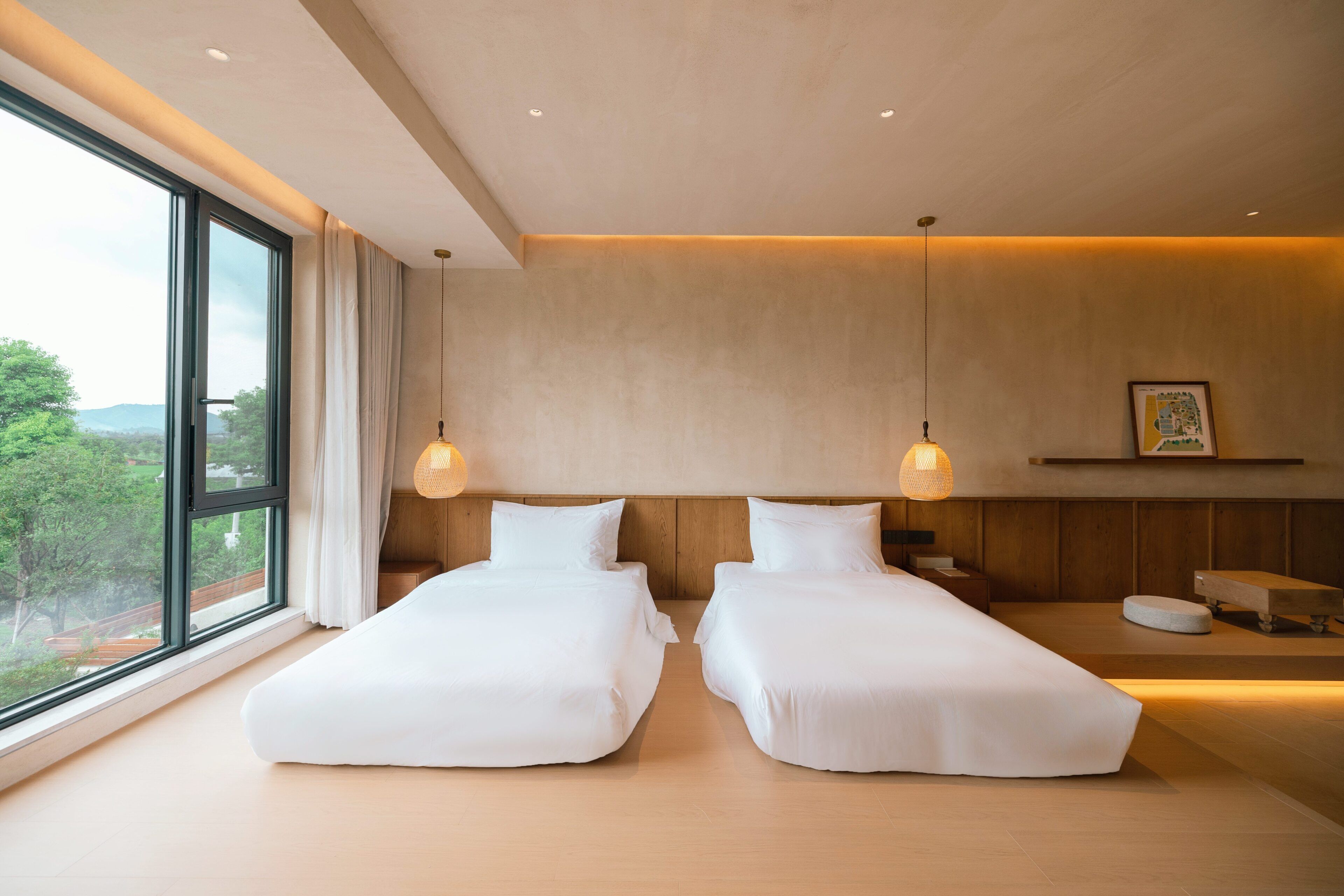 A modern hotel room with two beds, a large window, and a view of greenery.