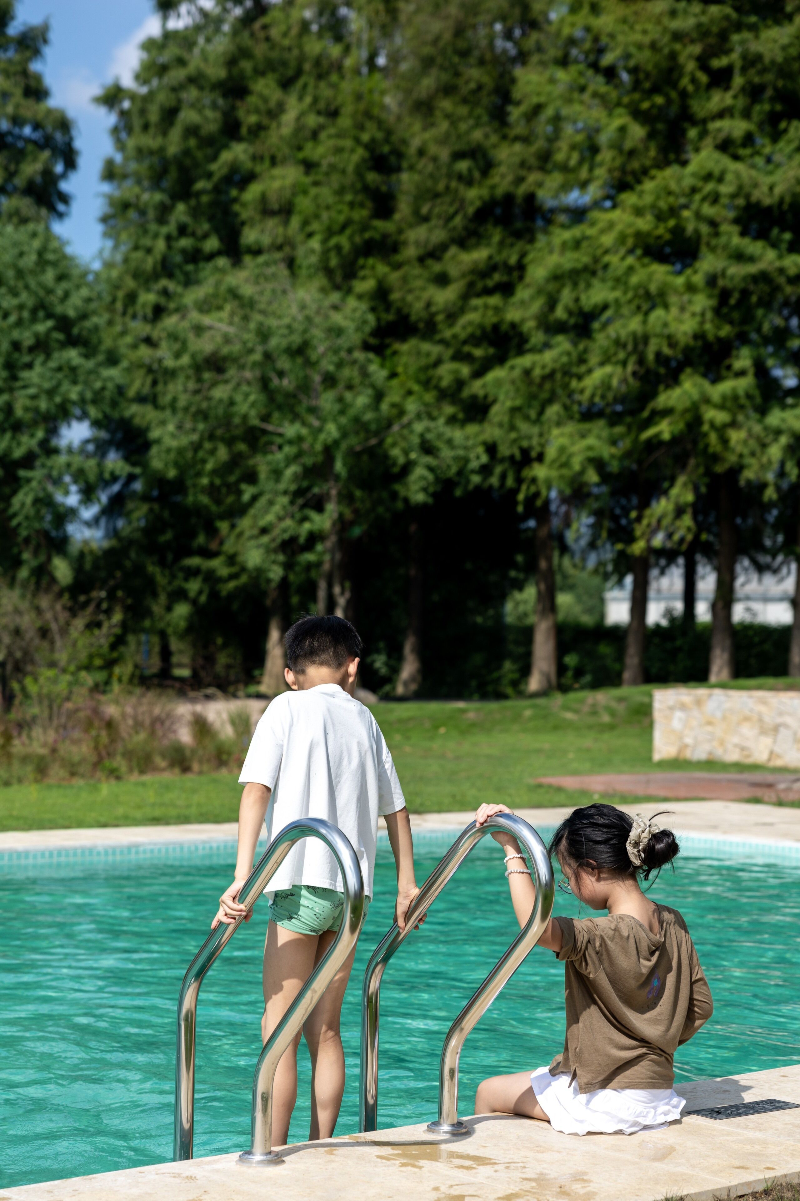 A poolside area with metal pool ladders, a stone wall, and a grassy area with trees in the background.