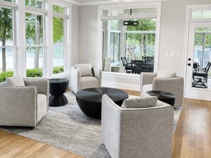 Een moderne woonkamer met grote ramen, een zwart, rond poef en grijze gestoffeerde fauteuils.