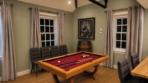 A pool table with balls, chairs, and a wooden barrel in a room with windows and curtains.
