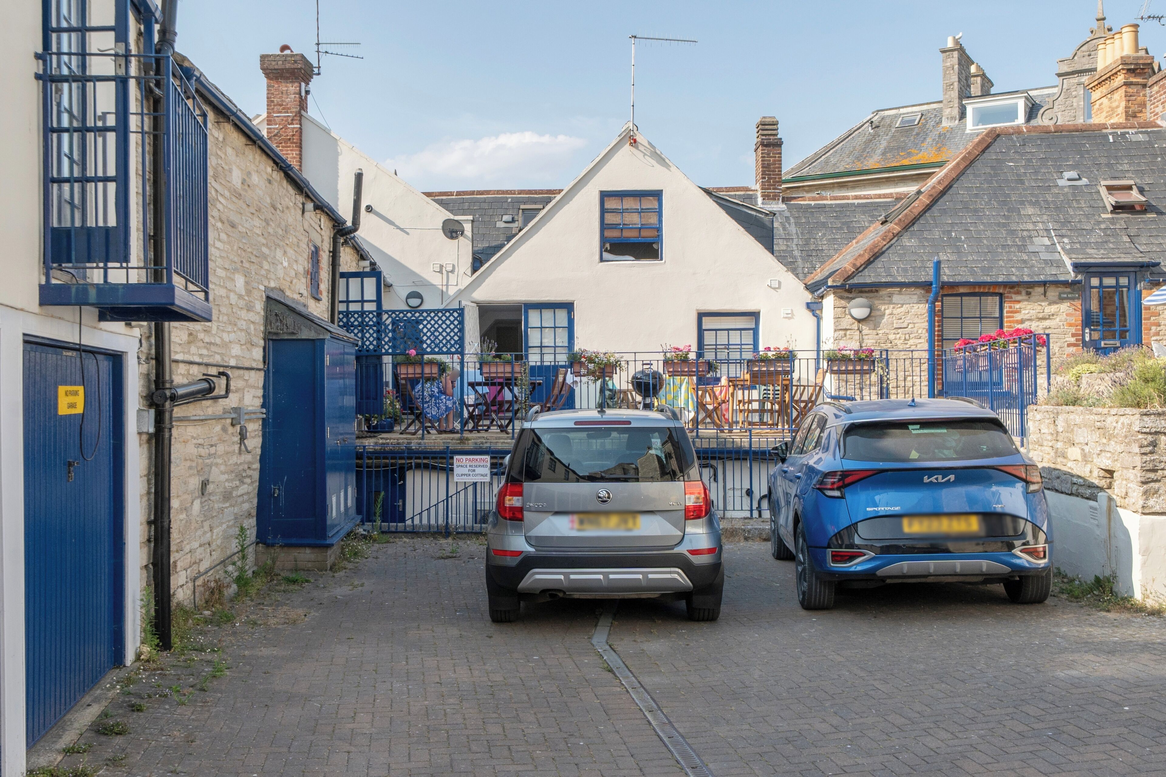 A narrow street with two cars parked on the side, a building with a white facade and blue shutters, and a blue gate with a 'NO PARKING' sign.