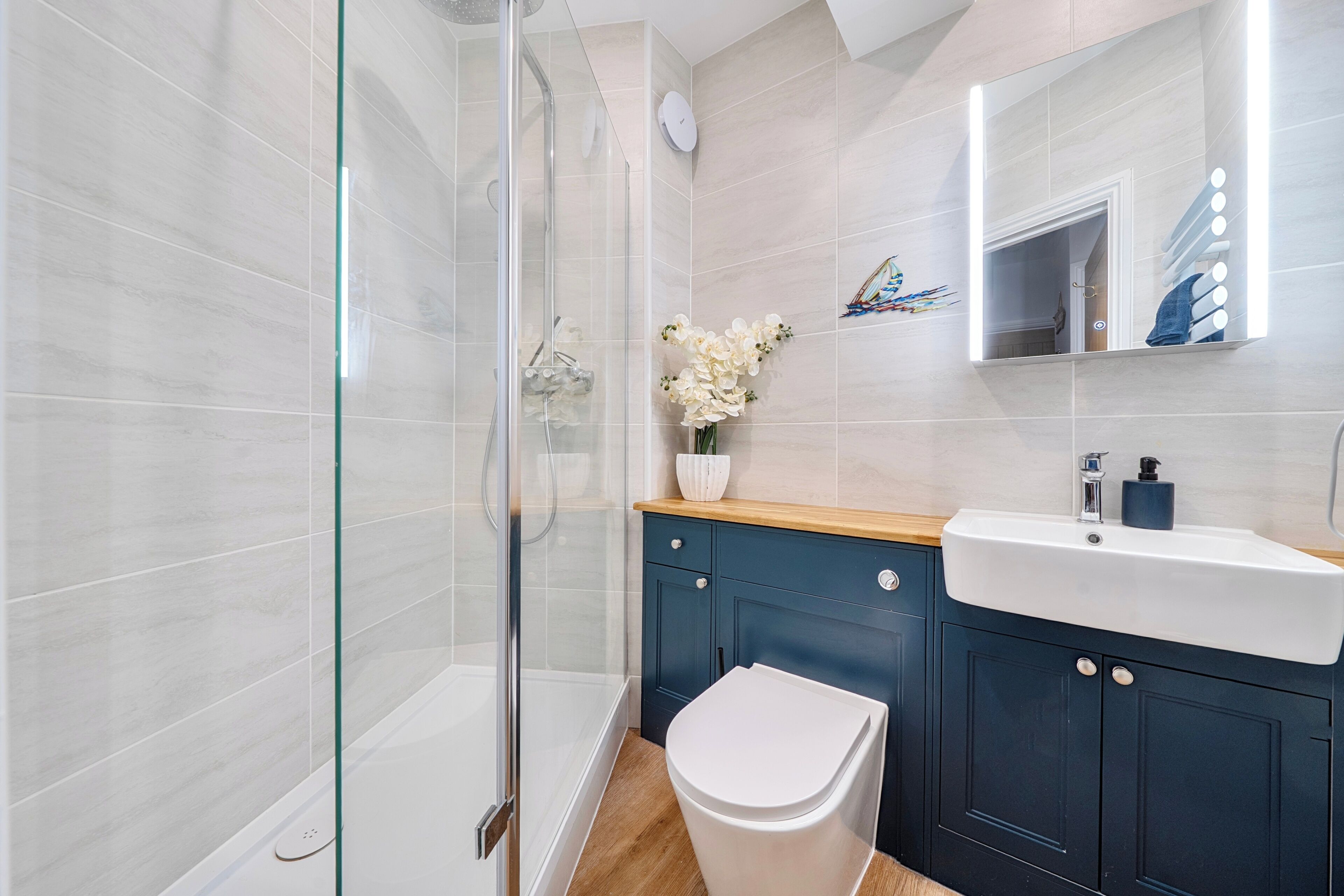 Une salle de bain moderne avec une douche à l’italienne en verre, des toilettes blanches, un meuble-lavabo en bois et un miroir avec éclairage intégré.
