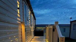 Una escalera de madera conduce a un porche cubierto con luces colgantes, junto a una casa rodante.