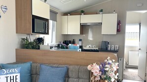 Una sala de estar moderna con un sofá gris, una pequeña mesa de madera con un florero, y una cocina con armarios blancos y un microondas.
