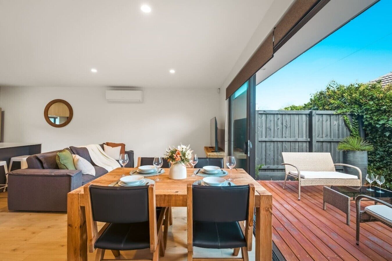 Un comedor moderno con una mesa de madera dispuesta para cuatro personas, un sofá y una silla de mimbre en una terraza de madera.