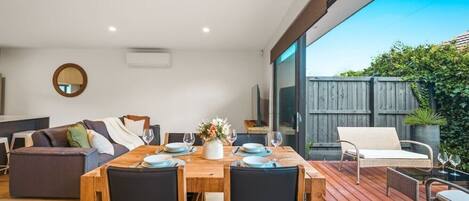 Un comedor moderno con una mesa de madera dispuesta para cuatro personas, un sofá y una silla de mimbre en una terraza de madera.